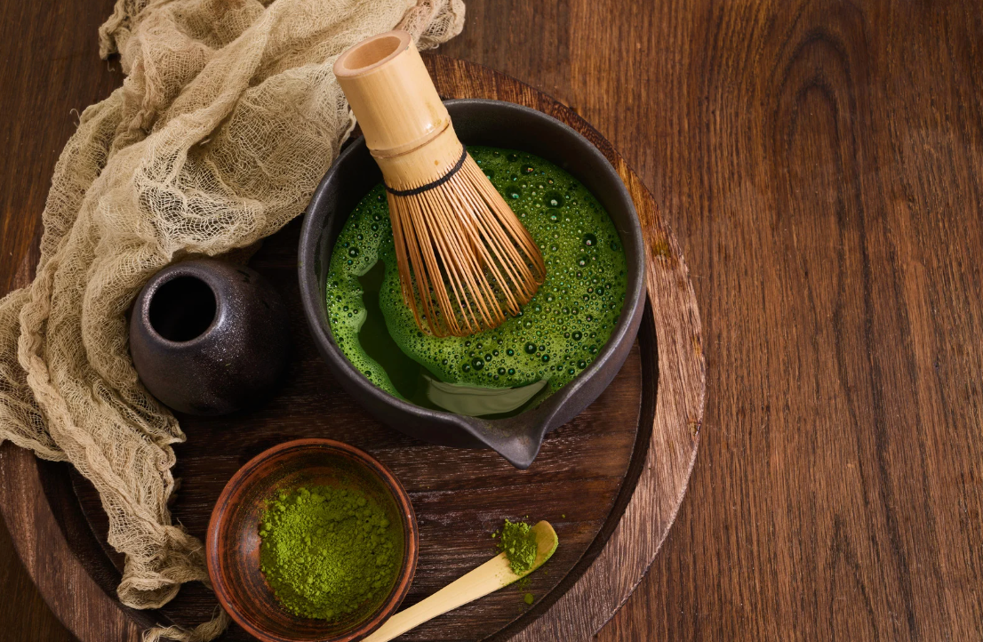 深色陶碗中的鮮綠抹茶配竹製茶筅，置於木質桌面上，旁邊有米色紗布與小陶瓶，右側留有大面積留白。
