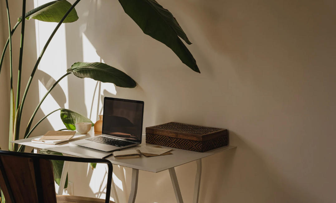 陽光灑進室內工作角落，桌上放有手提電腦、筆記本與植物，牆面留有大片暖色留白。