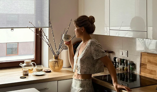 一名女子站在廚房窗邊喝茶，晨光照進室內，檯面放有玻璃茶壺、花枝與香料罐。