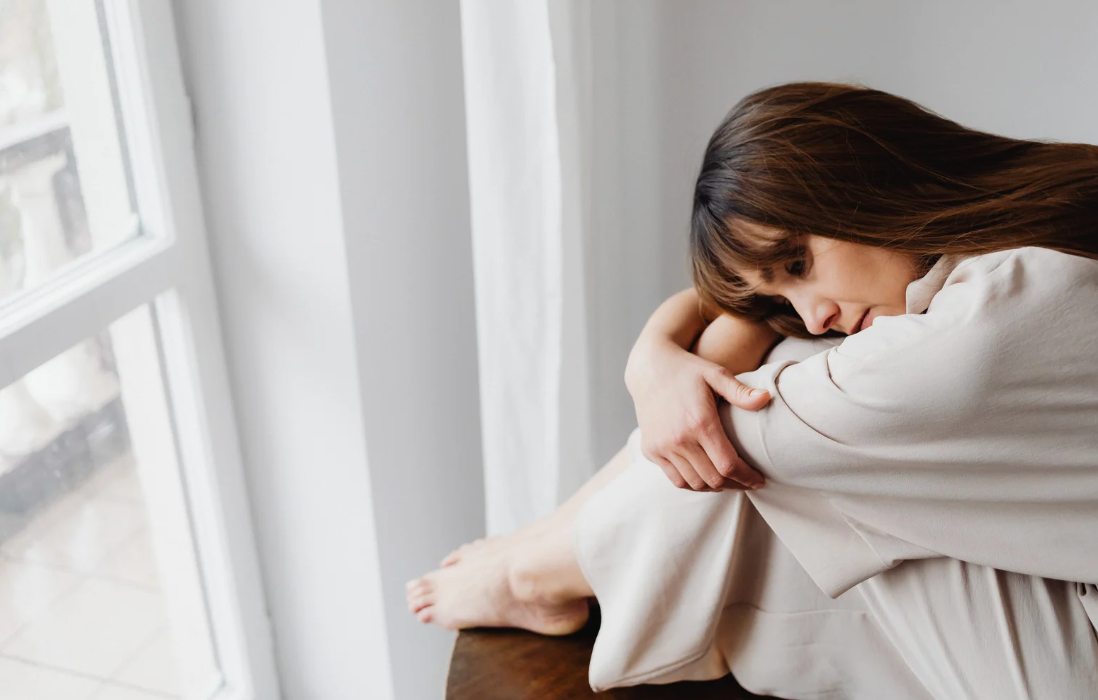一名女子抱膝坐在窗前，下巴輕靠膝上，安靜沉思，呈現城市疲憊與情緒耗損的氛圍
