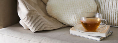 溫暖燈光下的透明玻璃杯茶放在書本上，旁邊是米色抱枕與沙發，呈現放鬆的晚間居家氛圍，象徵調整生活節奏與舒緩胃酸倒流的不適。